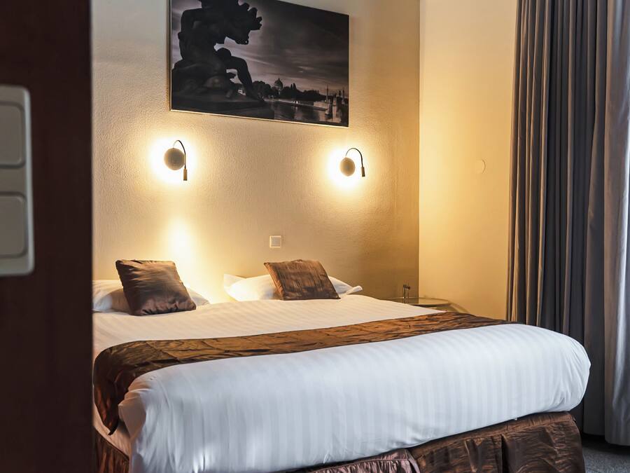 A hotel room with a large bed, two wall-mounted lights, a painting, and a window with curtains.