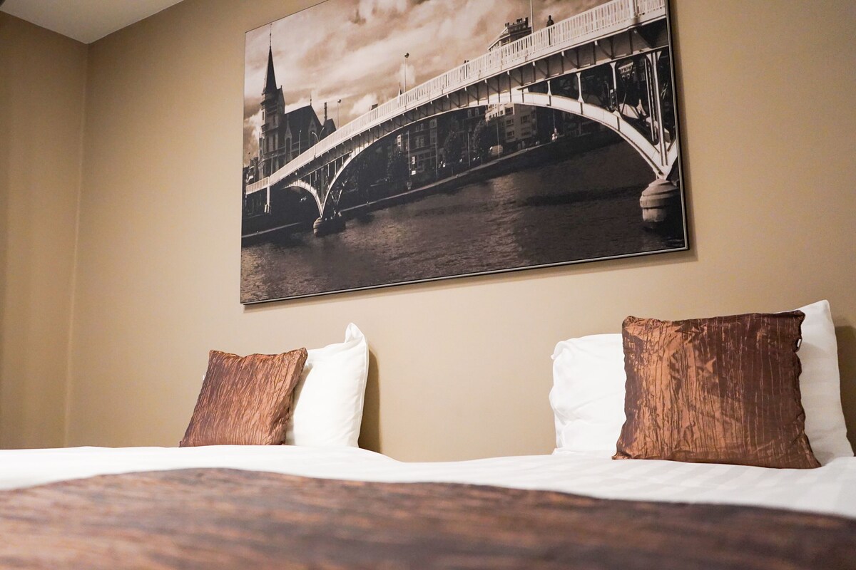 A hotel room with a bed, two pillows, a large framed picture of a bridge, and a curtain.