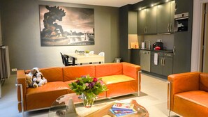 A modern living room with a sofa, a coffee table with books, a painting on the wall, and a kitchen area with cabinets and appliances.