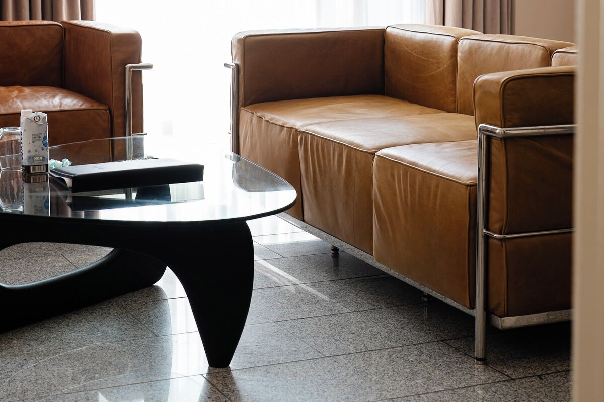 A modern living room with a brown leather sofa, a glass coffee table, and a view through sheer curtains.