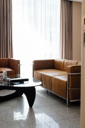 Un salón moderno con un sofá de cuero marrón, una mesa de centro de vidrio y vistas a través de cortinas traslúcidas.