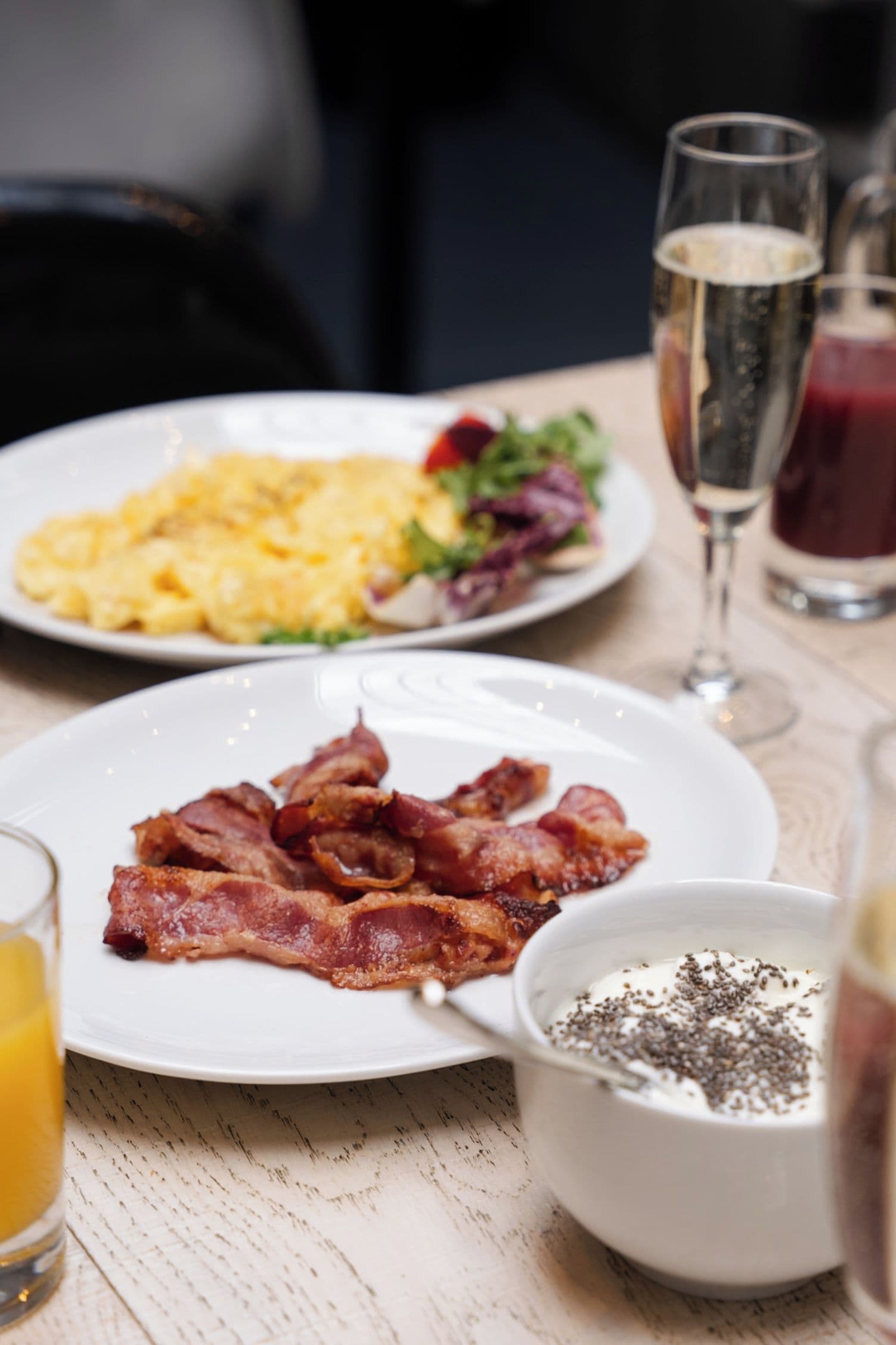 Una escena de desayuno con platos de tocino, huevos revueltos y una ensalada, junto con un vaso de jugo de naranja y un vaso de bebida roja.