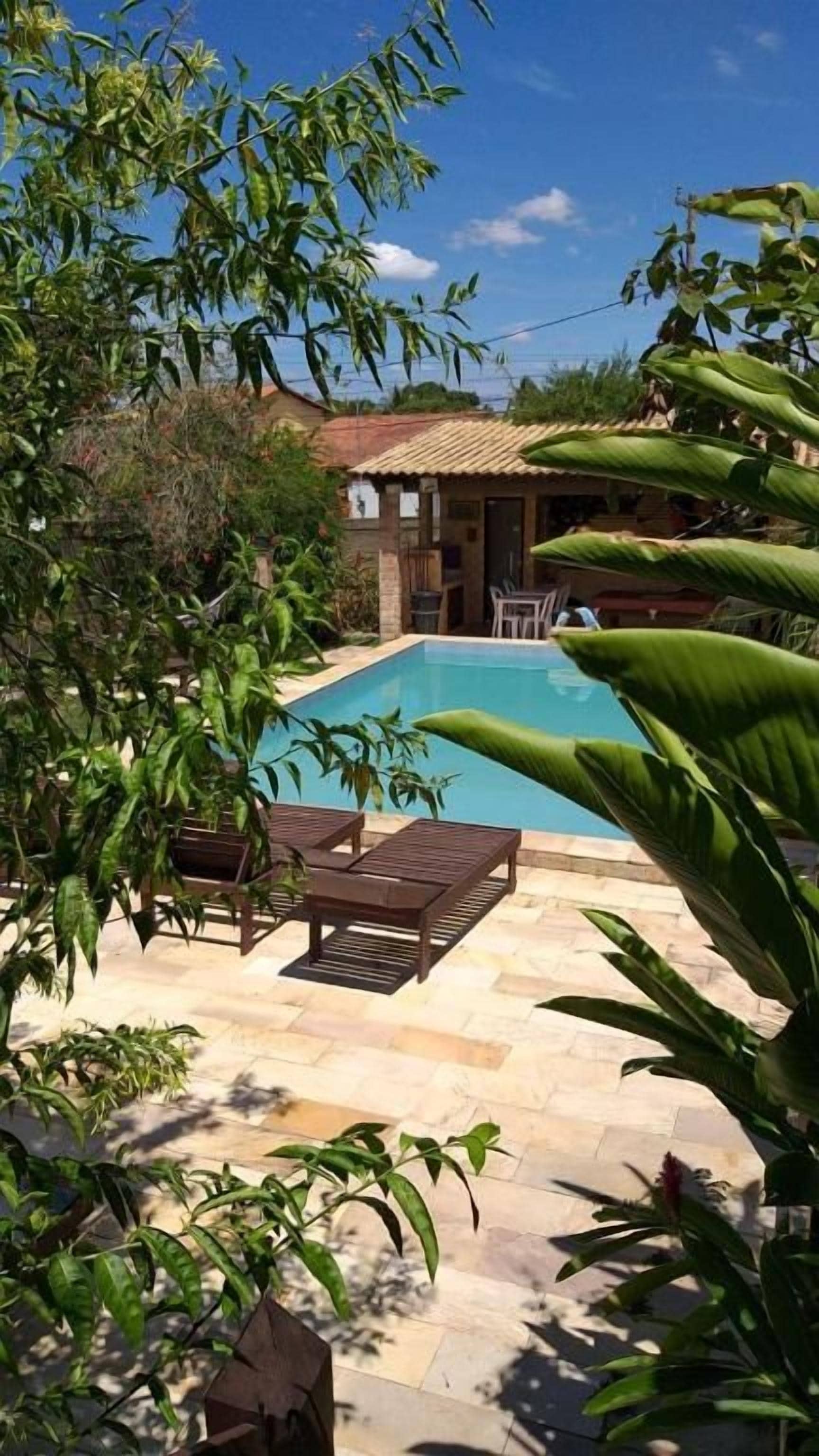 A swimming pool with a patio area, surrounded by greenery and a building with a tiled roof.