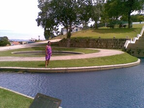A woman in a purple dress standing by a circular water feature with a stone wall and a grassy area. - Recanto Suítes (Araruama)