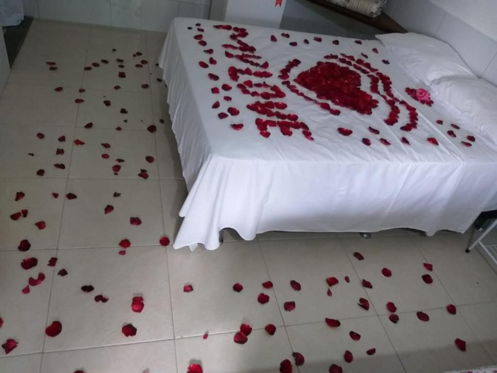 A bed with a white bedspread and a heart-shaped arrangement of red rose petals on the floor.