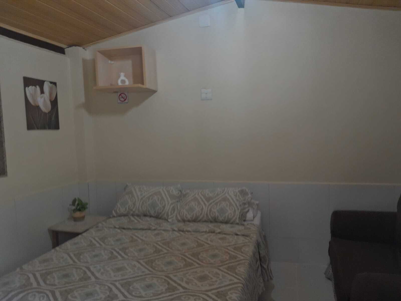 Ein Schlafzimmer mit einem Bett, einem kleinen Nachttisch mit Pflanze, einem wandmontierten Regal mit Vase und einem gerahmten Bild.