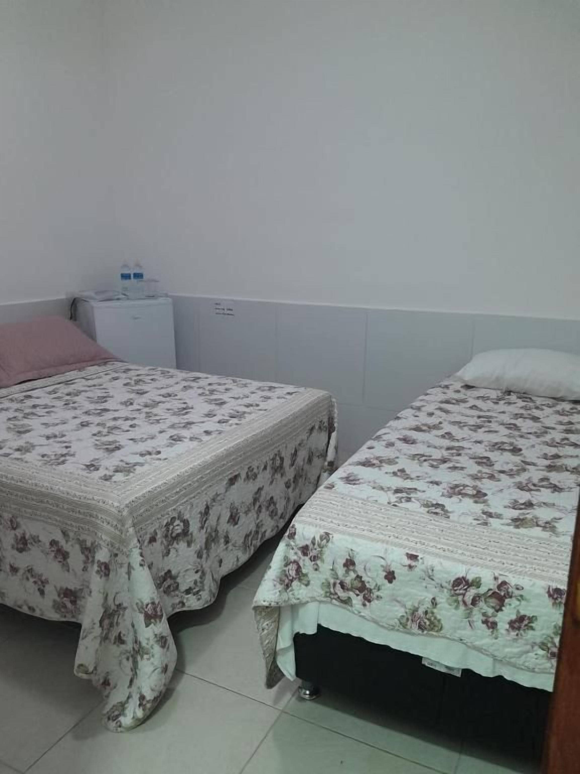 Dos camas individuales con ropa de cama floral, una mesita de noche pequeña y un mueble de pared con dispensador de agua.