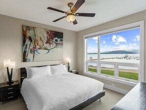 A bedroom with a large bed, a ceiling fan, a painting, and a view of a lake and dock through a large window.