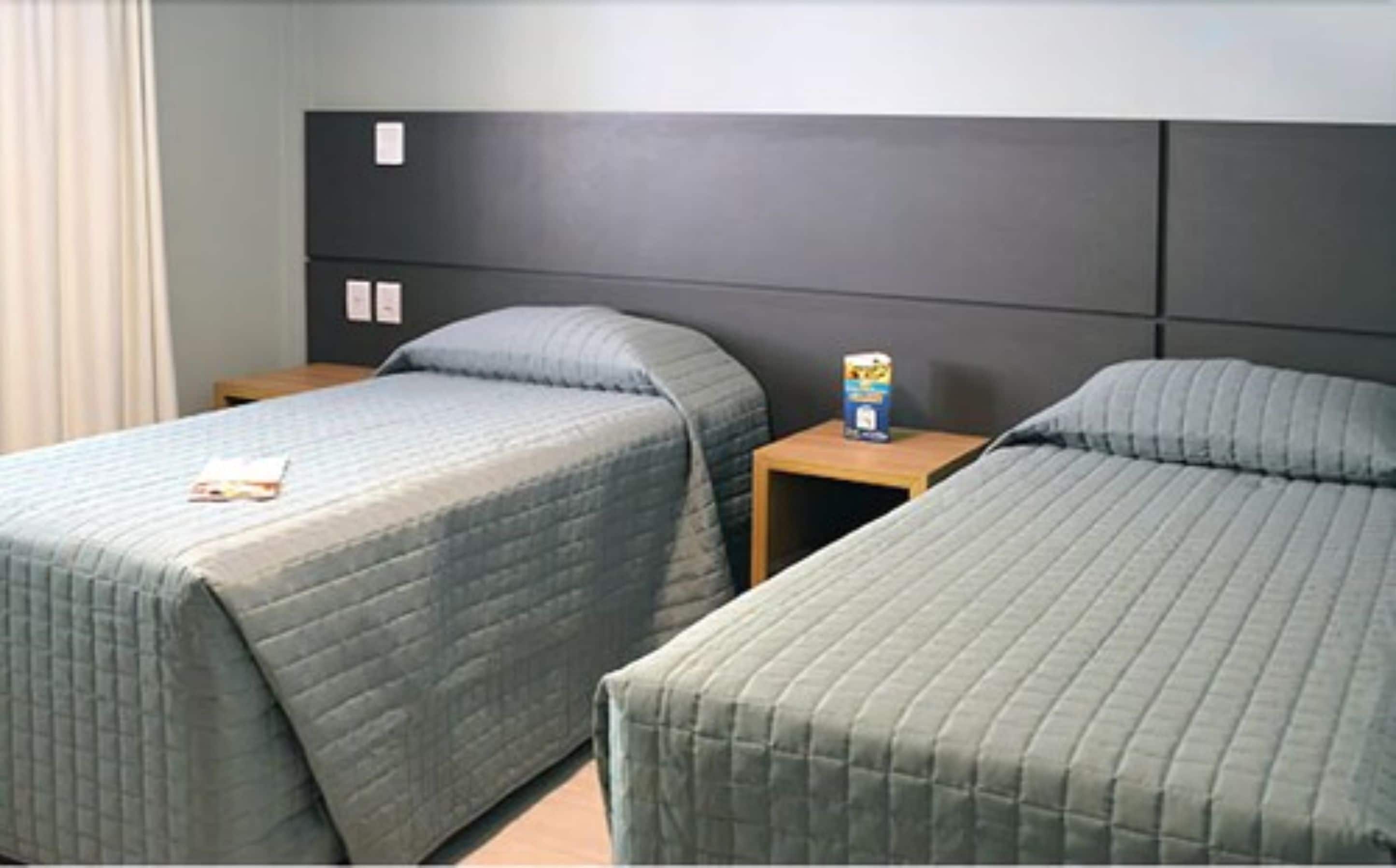 A hotel room with two single beds, a wooden nightstand with a book and a box, and a grey headboard.