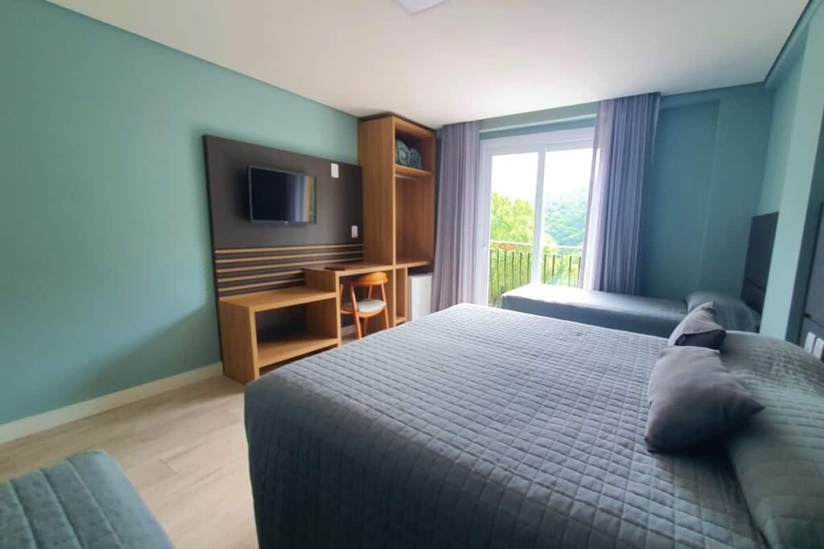 A bedroom with a bed, a TV mounted on the wall, a wooden desk, and a balcony with curtains.