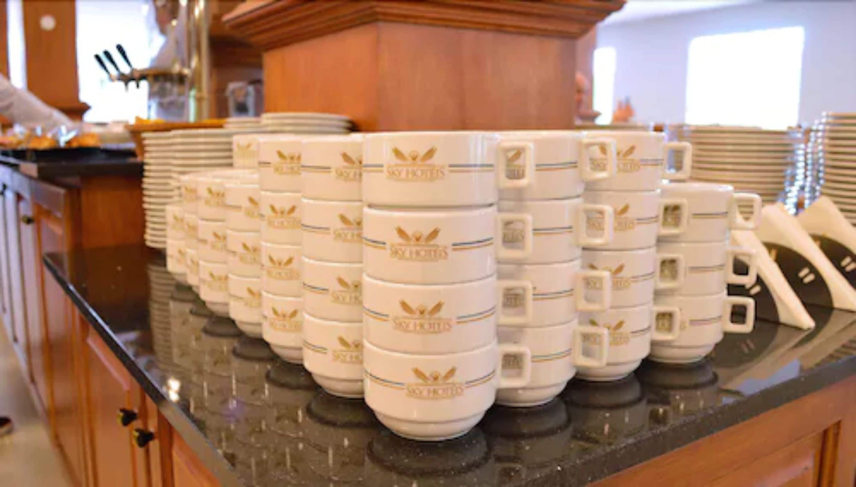 Stacks of white mugs with gold emblems on a kitchen counter.