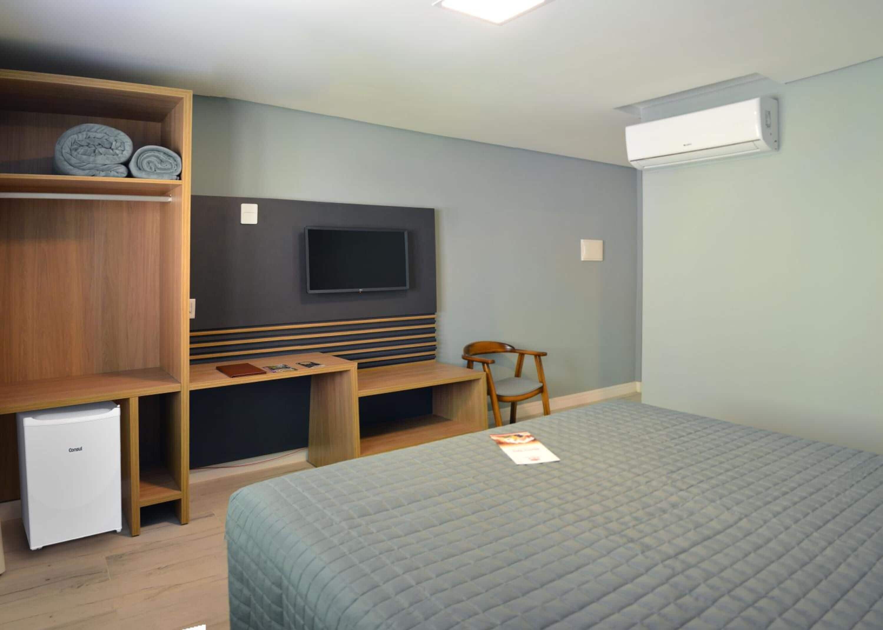 A bedroom with a bed, a desk, a chair, a TV, a wardrobe, and a small fridge.