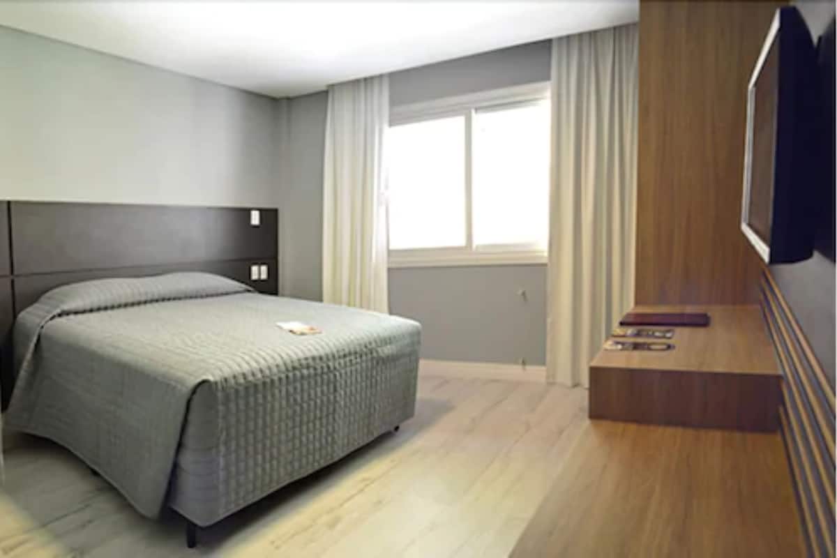A modern hotel room with a bed, a TV, and a window with curtains.
