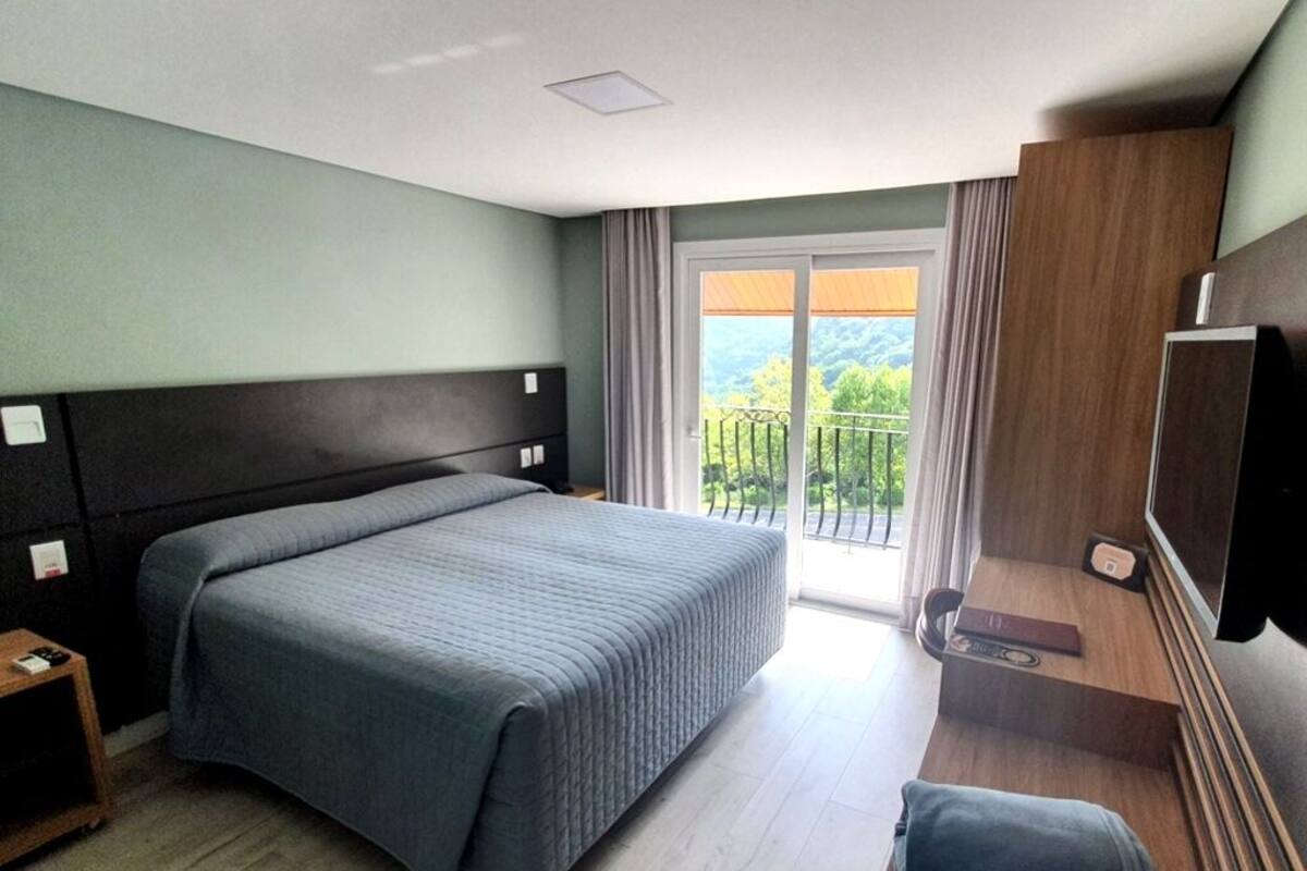 A hotel room with a bed, a TV, a small table, and a balcony with a view of greenery.