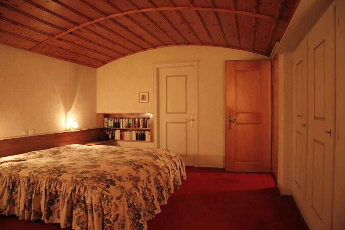 A bedroom with a wooden ceiling, a bed with a floral bedspread, a bookshelf, and a door.