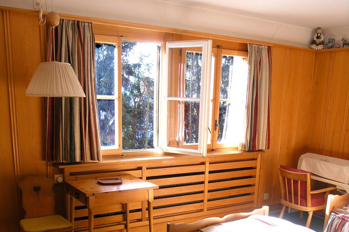 A cozy room with a bed, a desk, and a chair. There are windows with curtains, a rug, and a lamp.