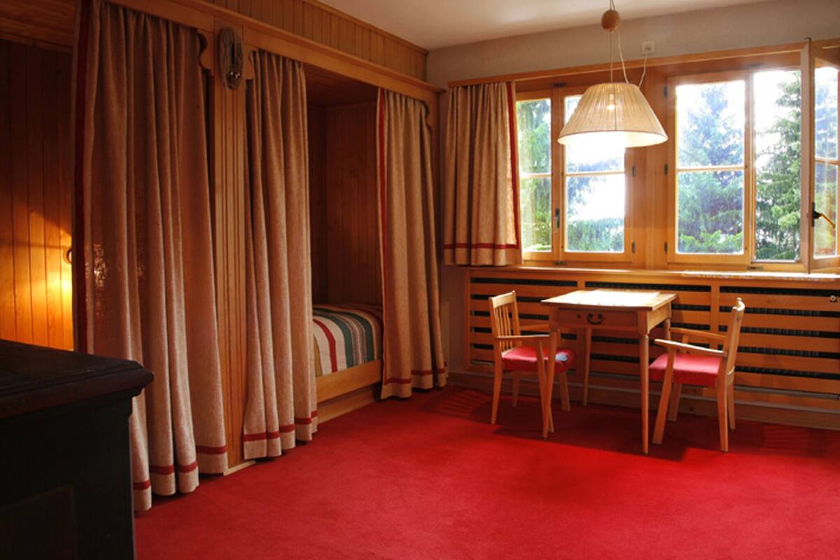 A cozy room with a bed, a desk, and two chairs. There are curtains on the windows and a lamp hanging from the ceiling.