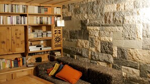 A cozy reading nook with a stone wall, wooden bookshelves, and a comfortable sofa with cushions. - B&B Arche de Noé (Bagnes)