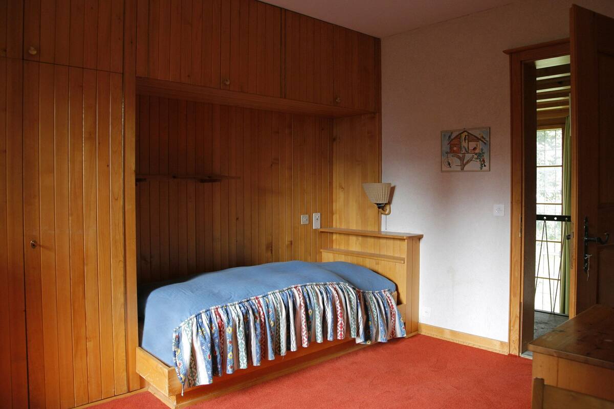 A bedroom with a bed, wooden walls, a small table with a lamp, and a door leading to another room.