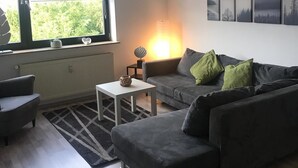 A modern living room with a grey sofa, a white coffee table, and a floor lamp. There is a large window providing natural light and a view of greenery outside. - Ferienwohnungen AKH Home (Sankt Wendel)