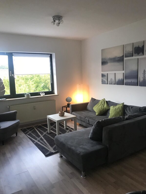 A modern living room with a grey sofa, a white coffee table, and a floor lamp. There is a large window providing natural light and a view of greenery outside. - Ferienwohnungen AKH Home (Sankt Wendel)