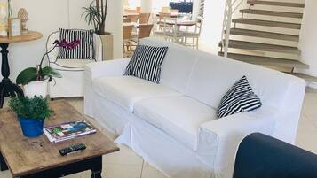 A spacious living area with a white sofa, a coffee table, and a blue sofa. There is a staircase in the background leading to an upper level.