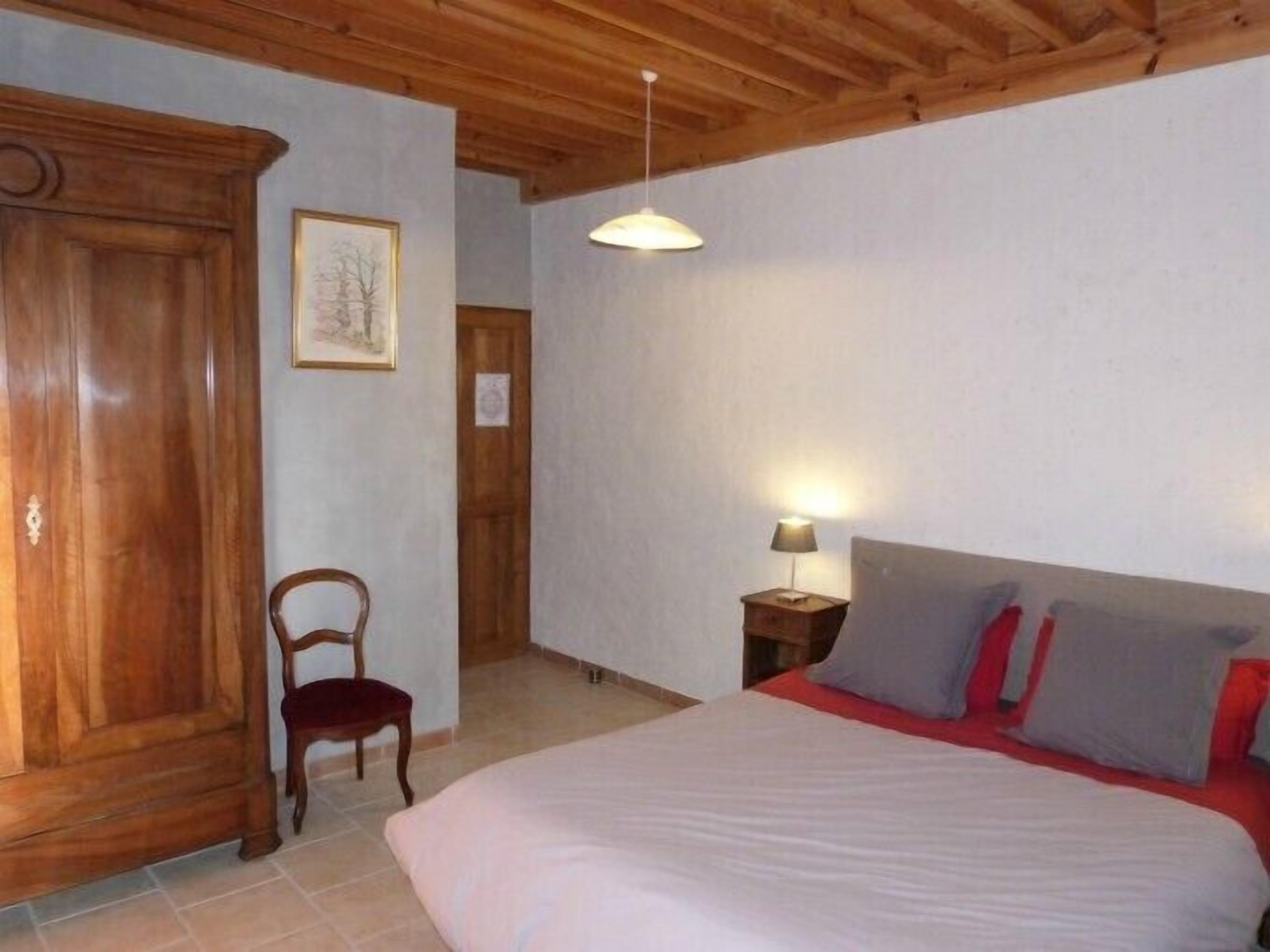 A bedroom with a wooden wardrobe, a single bed with red and grey pillows, a bedside table with a lamp, and a framed picture on the wall.