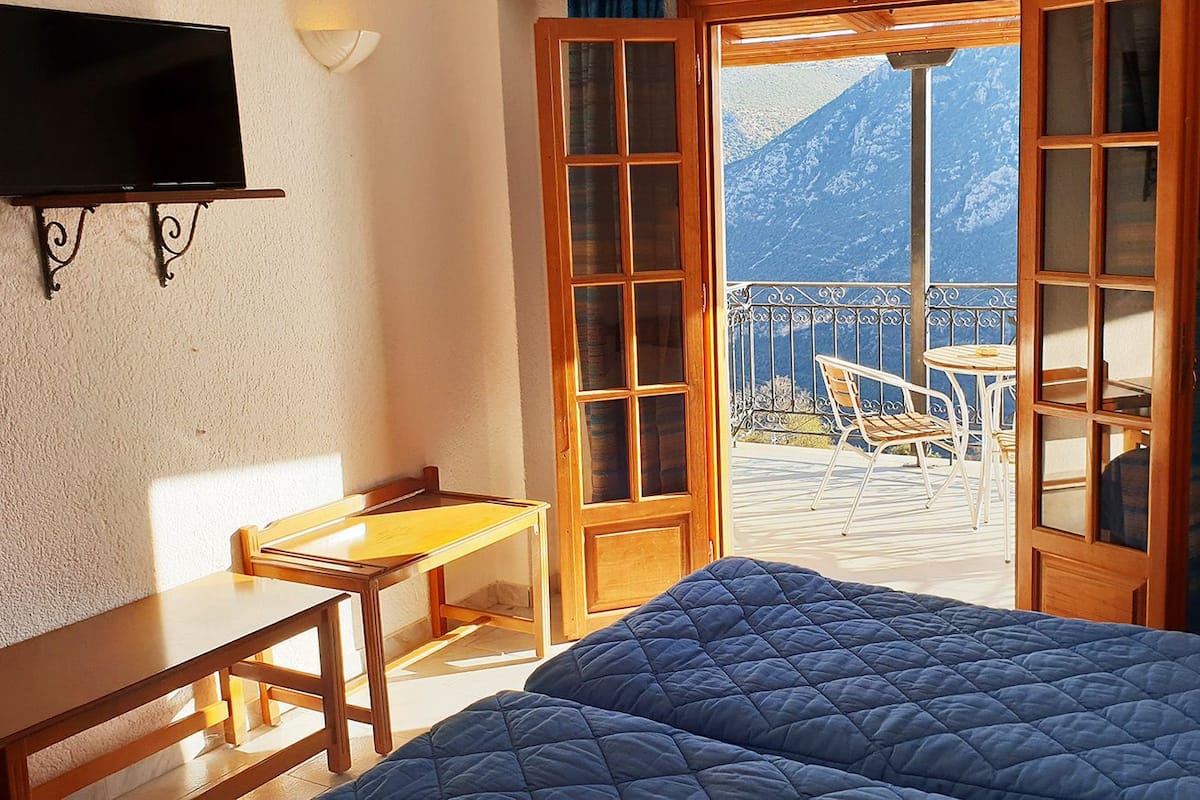 Un dormitorio con cama, escritorio, silla y televisor. Cuenta con un balcón con mesa y sillas.