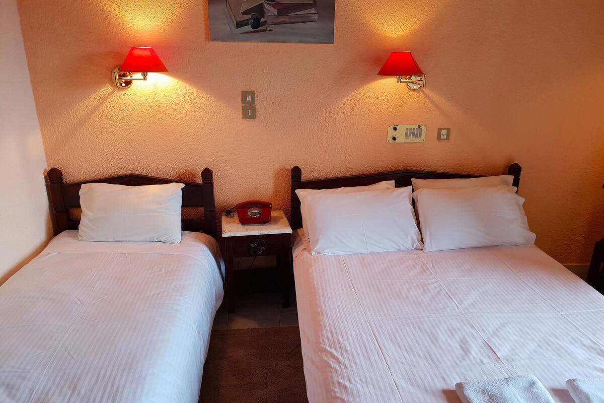 Habitación de hotel con dos camas individuales, cada una con sábanas y almohadas blancas, una mesita de noche con teléfono rojo, y lámparas fijadas a la pared.