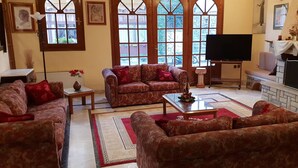 A living room with patterned sofas, a coffee table, a flat-screen TV, a fireplace, and a large window with a view. - Hotel Apollo (Delphi)