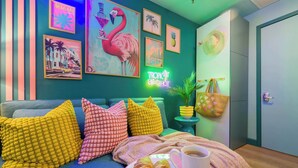 A cozy room with a teal wall, vibrant artwork, a teal sofa with colorful cushions, a small side table with a plant, and a plate of croissants and a cup of coffee. - ‼️Stylish Oceanfront‼️ Stay on Iconic Ocean Drive (Miami Beach)