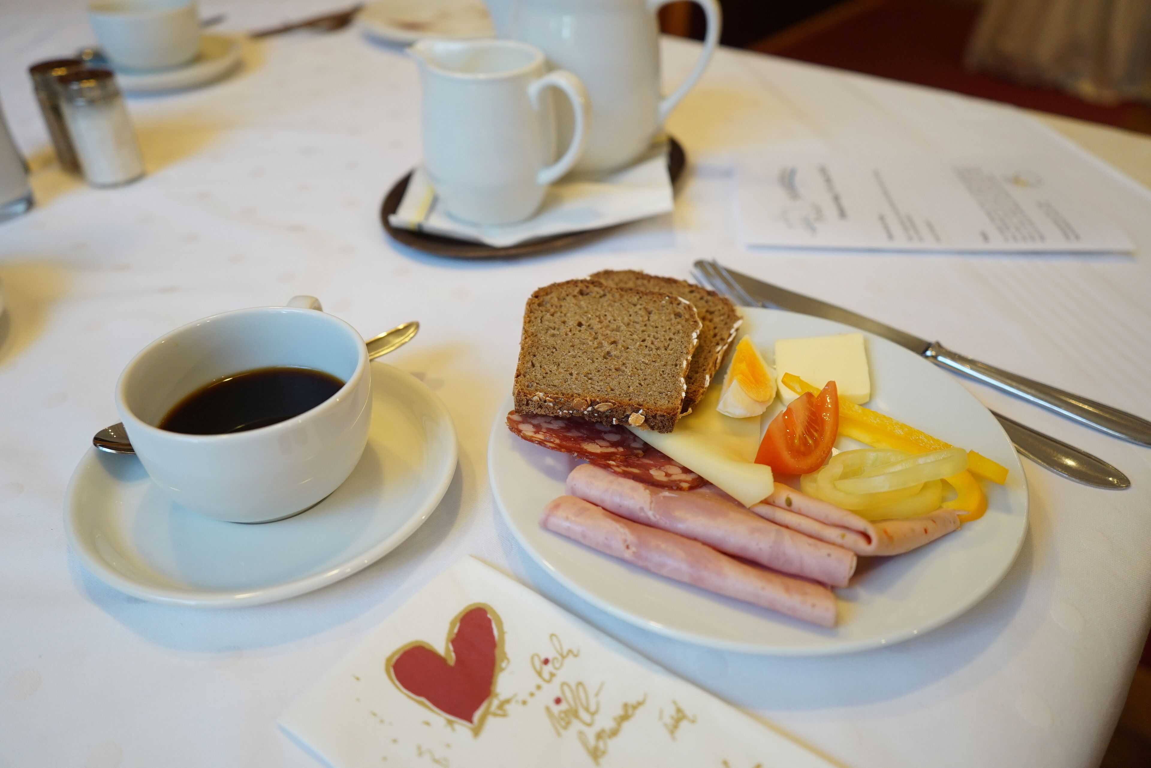 A breakfast table with a cup of coffee, a plate of ham, cheese, and vegetables, and a heart-shaped card.