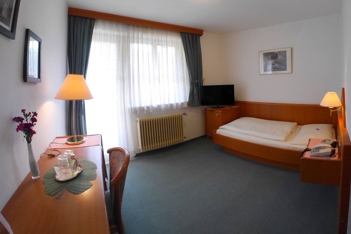 Habitación de hotel con cama, escritorio, silla, televisor, lámpara, radiador y ventana con cortinas.