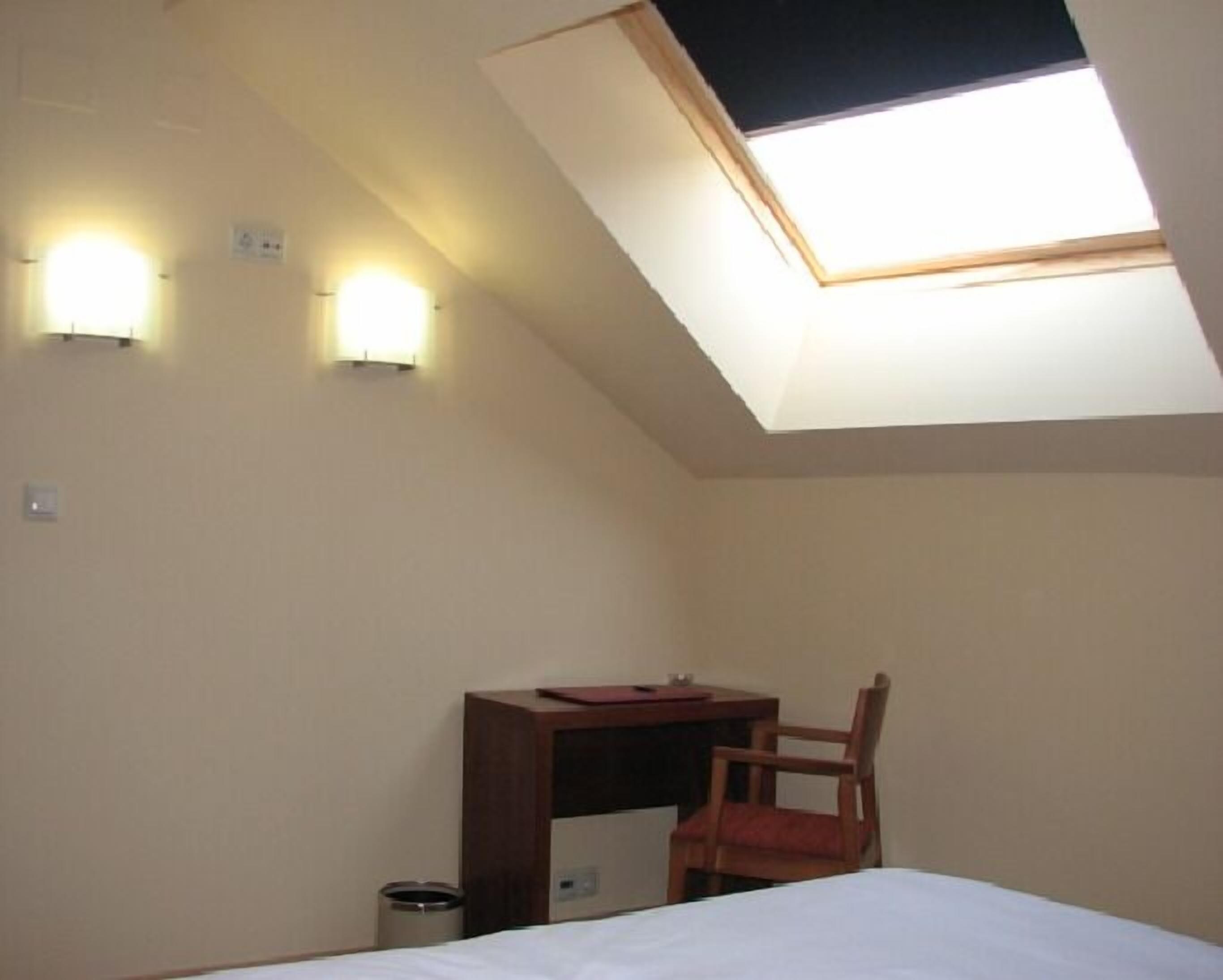 A small, simple room with a slanted ceiling, a bed, a desk, a chair, and two wall-mounted lights.