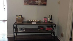 Una consola de madera con un cuadro abstracto rústico encima, una cesta con revistas, una botella de vino y algunos libros.