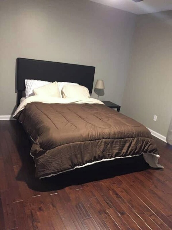 Un dormitorio con piso de madera oscura, una cama con cabecera oscura, almohadas blancas y una manta color beige.
