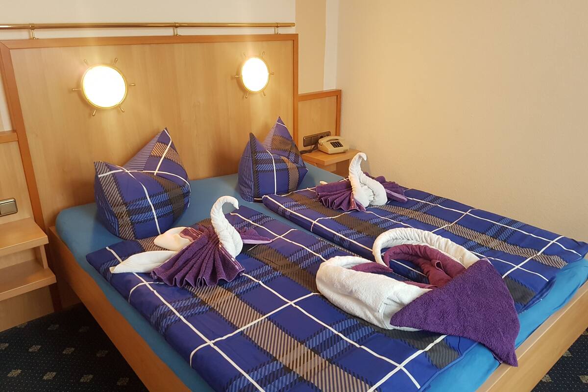 Habitación de hotel con cama, colcha a cuadros azul y blanco, dos almohadas, mesita de noche con lámpara y teléfono.