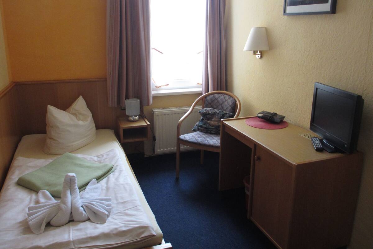 Habitación de hotel con cama, escritorio con computadora y televisor, una silla, lámpara y ventana con cortinas.