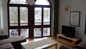 A living room with a large arched window, a television on a wooden stand, and a painting on the wall. - Haus Lucie (Heringsdorf)