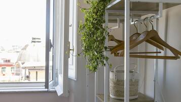 A white metal shelf with wooden hangers and a woven basket, next to a window with a view of buildings.