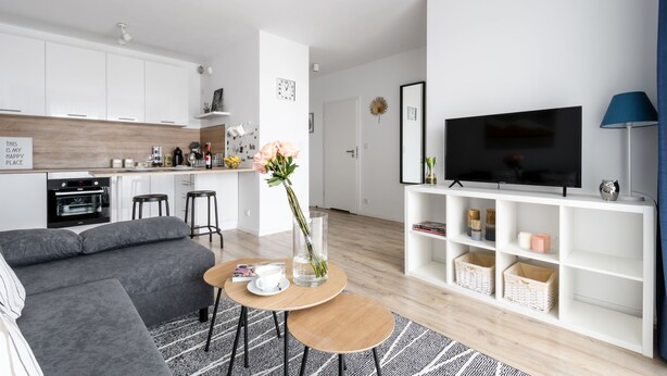 Una sala de estar moderna con un sofá gris, una mesa de centro, un televisor sobre un mueble blanco y una cocina al fondo.