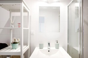 Une salle de bain moderne avec un lavabo blanc, deux miroirs, une douche en verre et une étagère sur laquelle est posé un vase à fleurs.