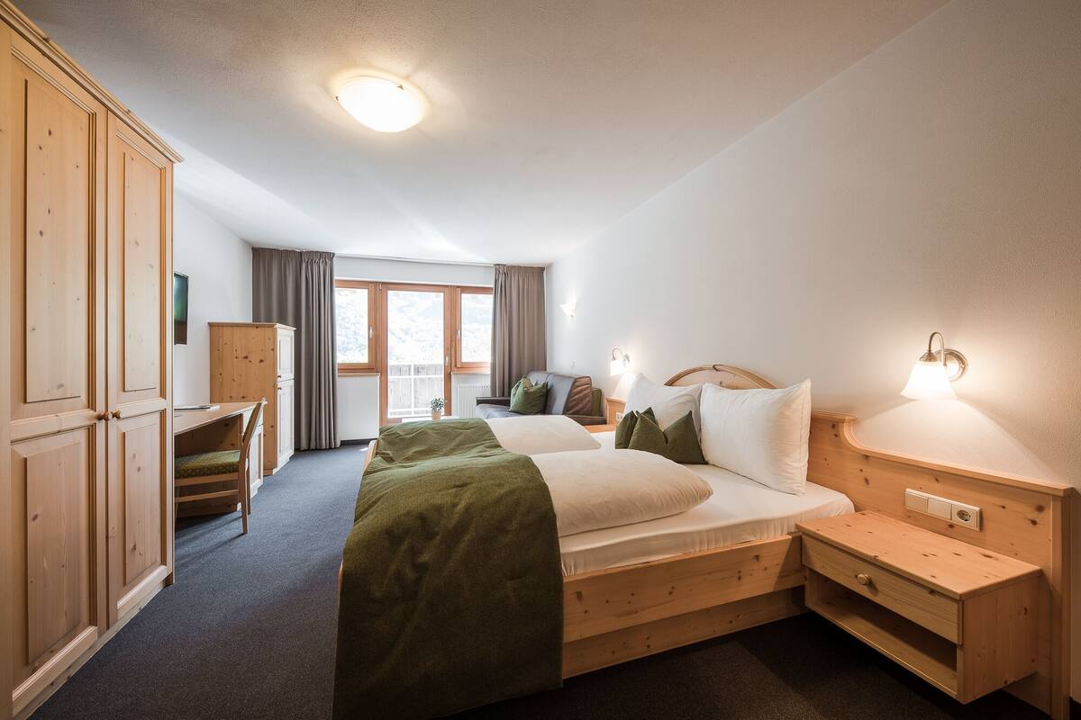 A hotel room with a large bed, a wooden wardrobe, a desk, and a window with curtains.