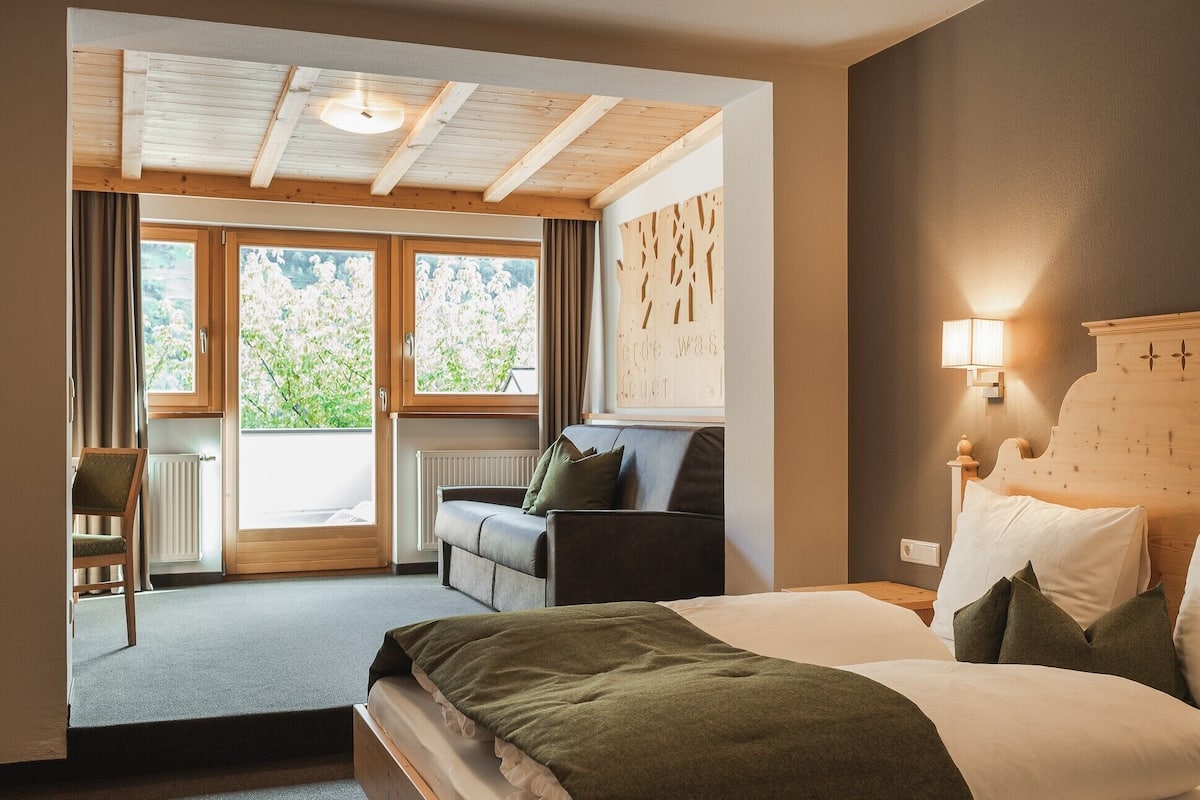 A bedroom with a bed, a chair, a sofa, a window, and a wall-mounted light fixture.