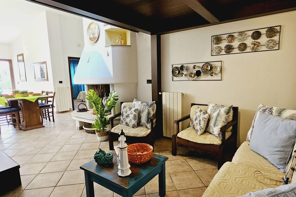 A living room with a sofa, chairs, a table, and decorative items. There is a dining area in the background with a table and chairs.