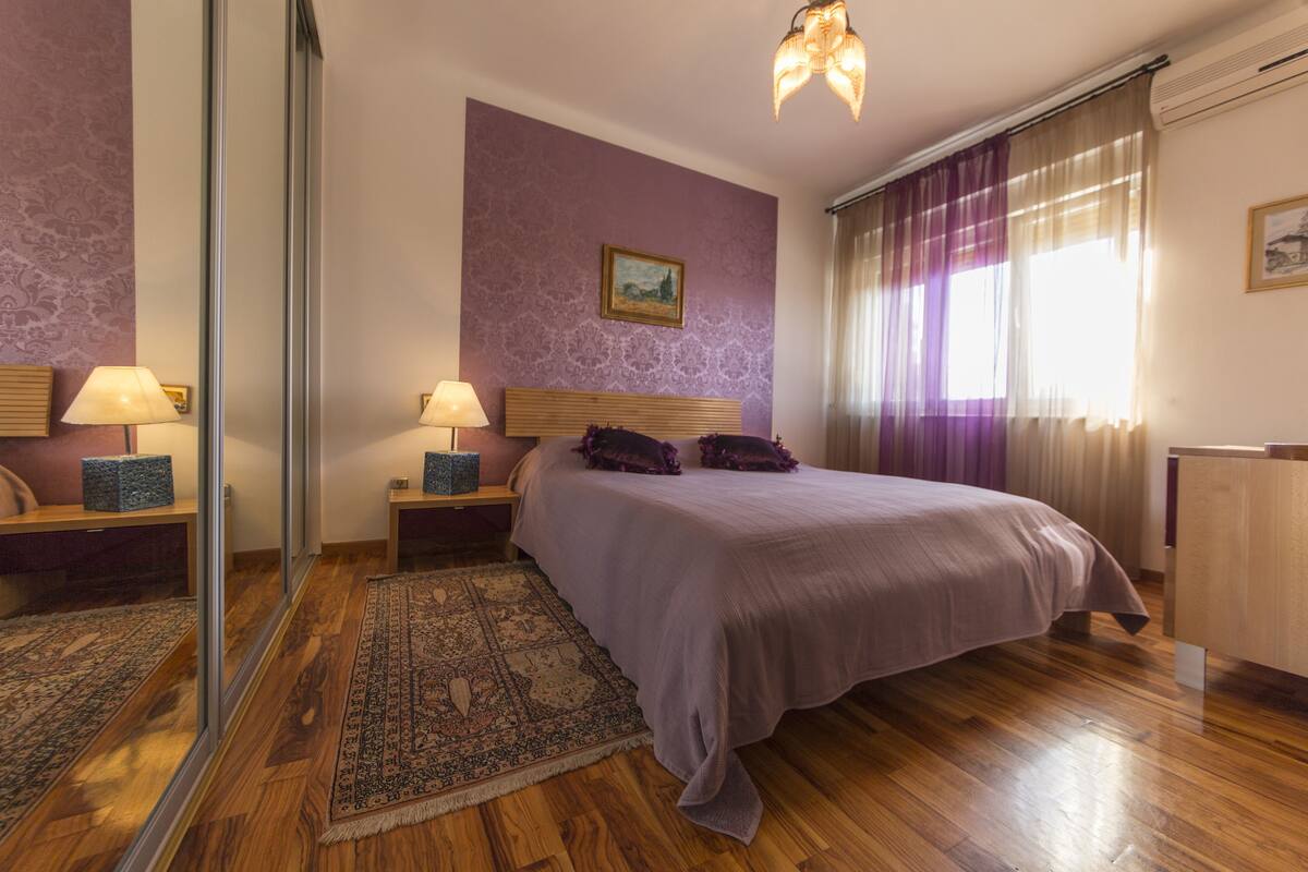 A bedroom with a large bed, wooden headboard, bedside tables with lamps, a rug, a painting on the wall, and a window with curtains.