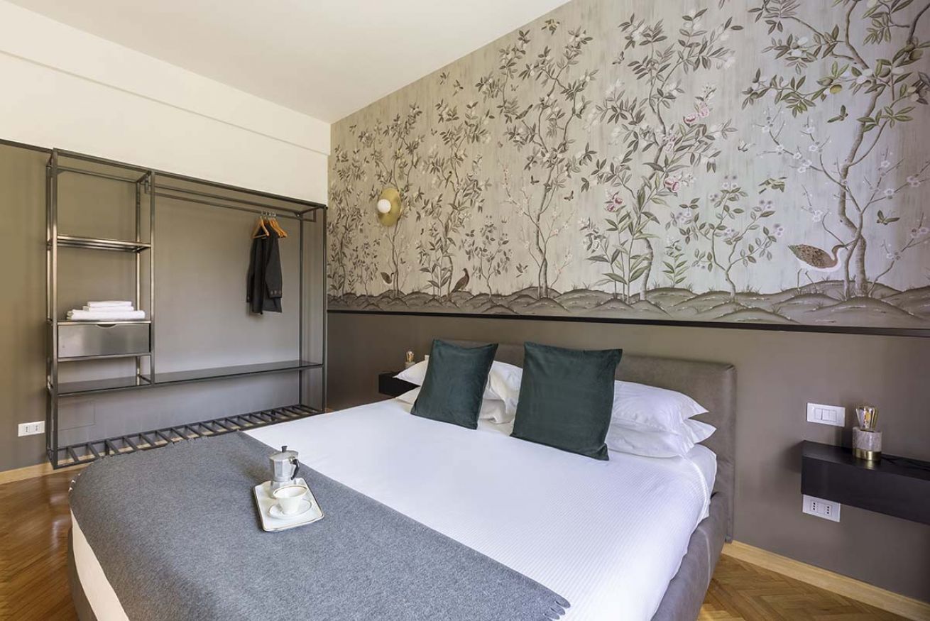 Una camera d'albergo con un letto, un armadio, uno scaffale con asciugamani, una lampada a parete e un affresco floreale.