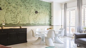 Un salon moderne avec un mur décoré d’un motif floral vert, des meubles blancs et une table basse en verre.
