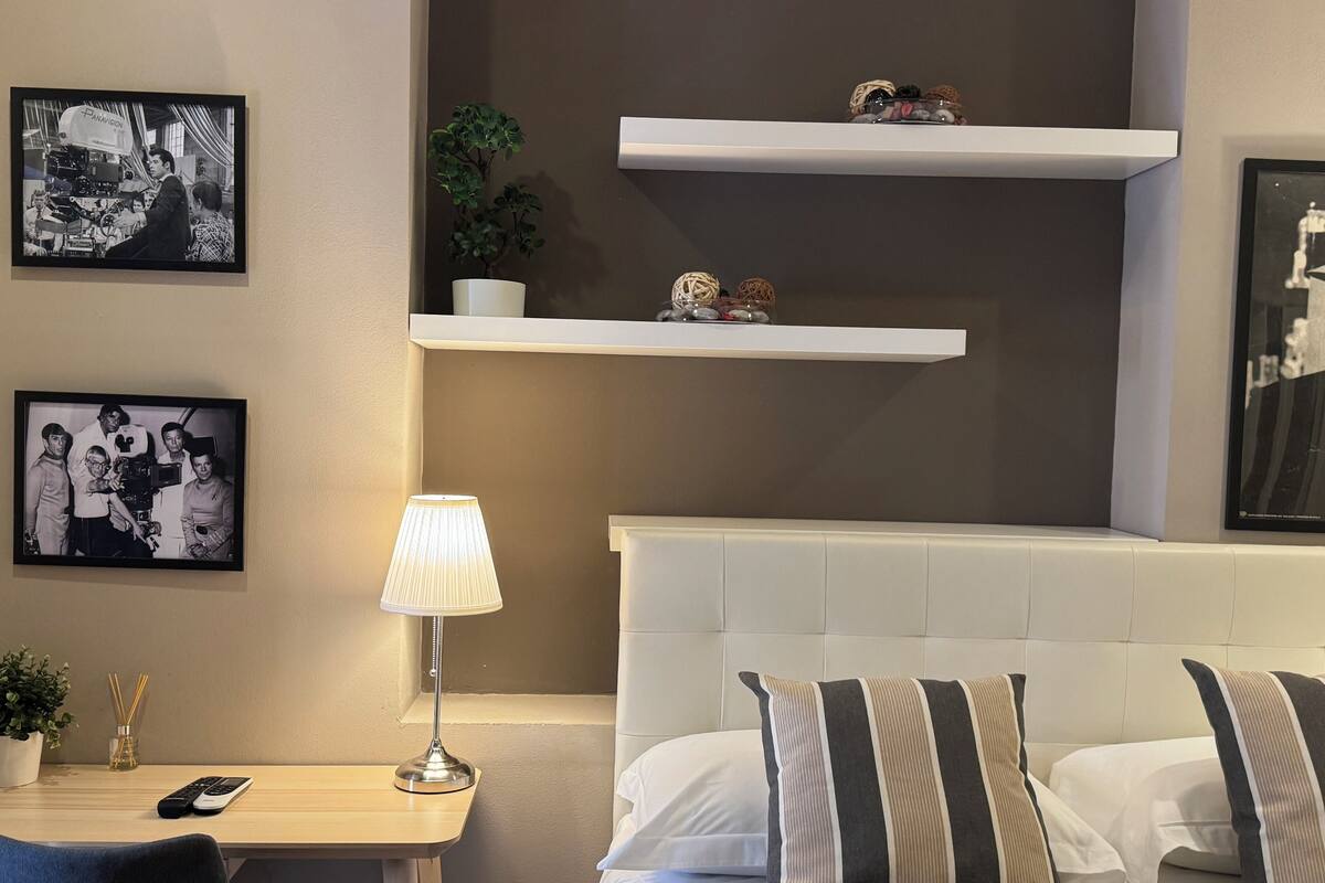 Een hotelkamer met een bed, een nachtkastje met een lamp, een klein bureau en twee planken met decoratieve objecten.