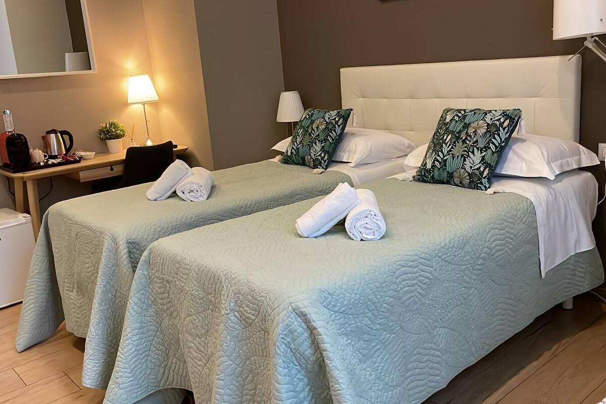 Een hotelkamer met twee bedden, elk voorzien van een lichtblauwe geborduurde spreidek en passende kussens. Er is een klein bureau met een lamp, een koffiezetapparaat en een waterkoker. Een witte verwarming is aan de linkerzijde van de kamer zichtbaar.